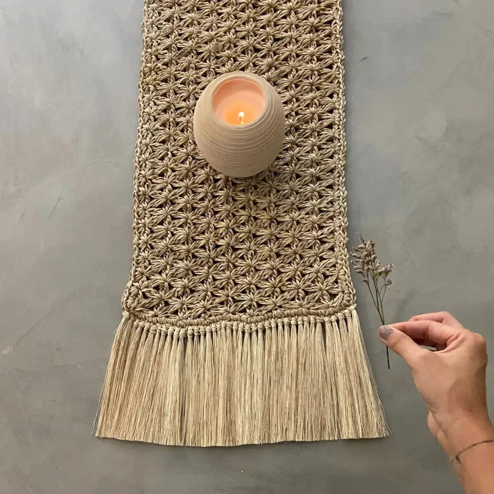 A handwoven table runner crafted from natural fique fibers with detailed knotting and a generous fringe. The close-up images show the rich texture and skillful craftsmanship. The runner sits beautifully on a rustic wooden dining table, paired with simple lit candle to highlight its organic, artisan-made appeal.